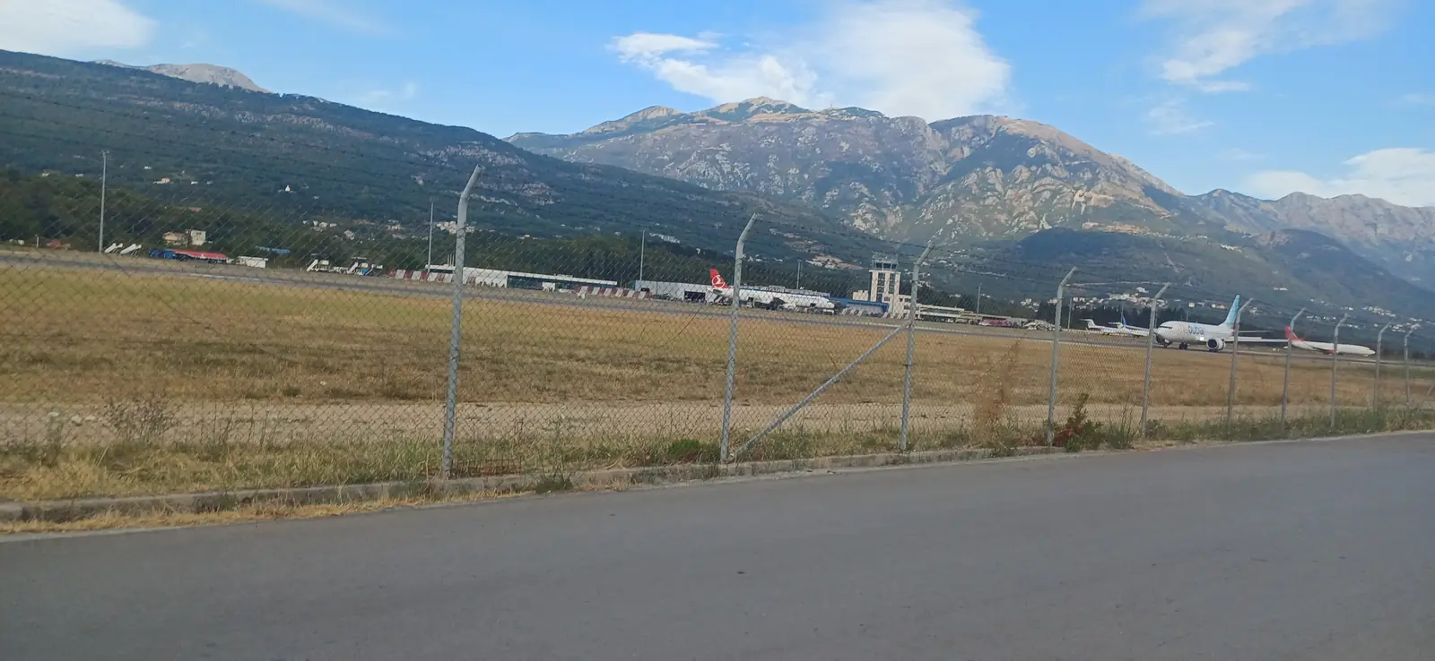 Silnice podél plotu letiště Tivat, za plotem stojící letadla a hory