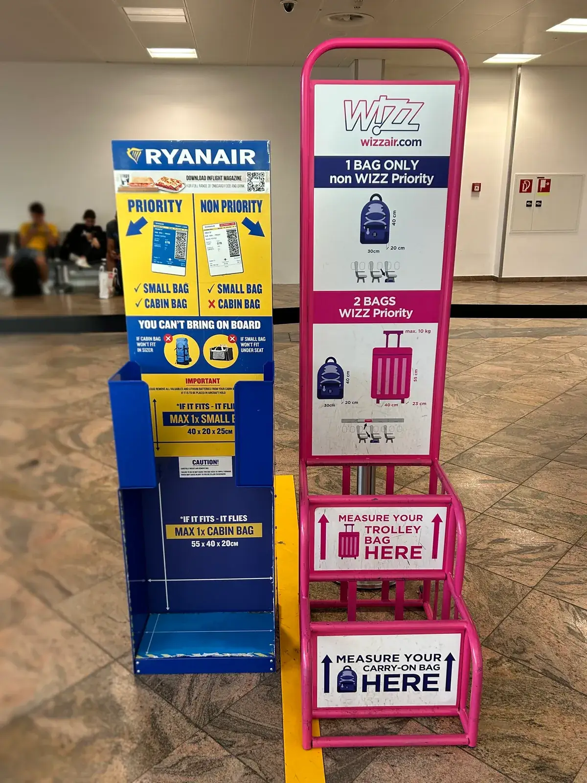 Modrý měřák zavazadel Ryanairu vedle růžového měřáku Wizz Airu v odletové hale